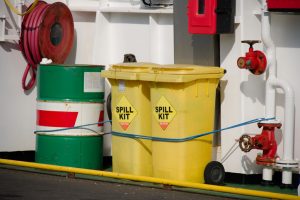 Yellow spill kit container on a ship deck beside green oil drums and red valves, with a blue rope securing items nearby.