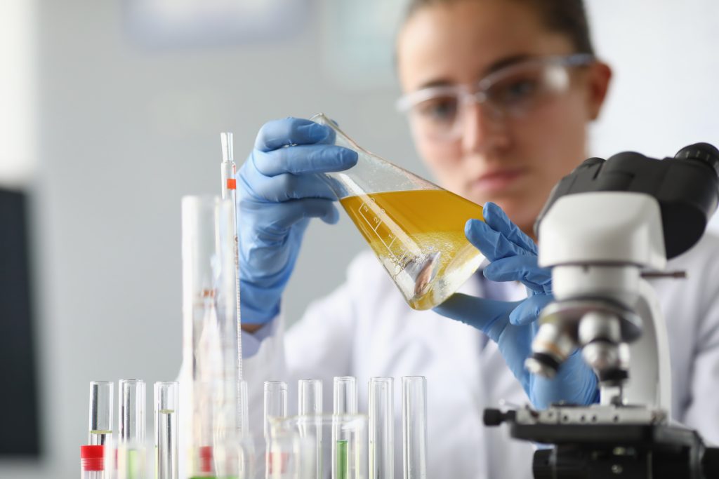 Scientist wearing blue gloves and safety goggles pours yellow liquid from a flask into a beaker in a lab.