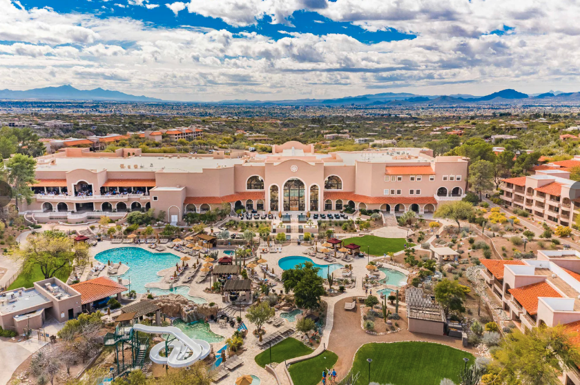 A large resort with pink stucco buildings, courtyards, and a sprawling pool complex viewed from above.