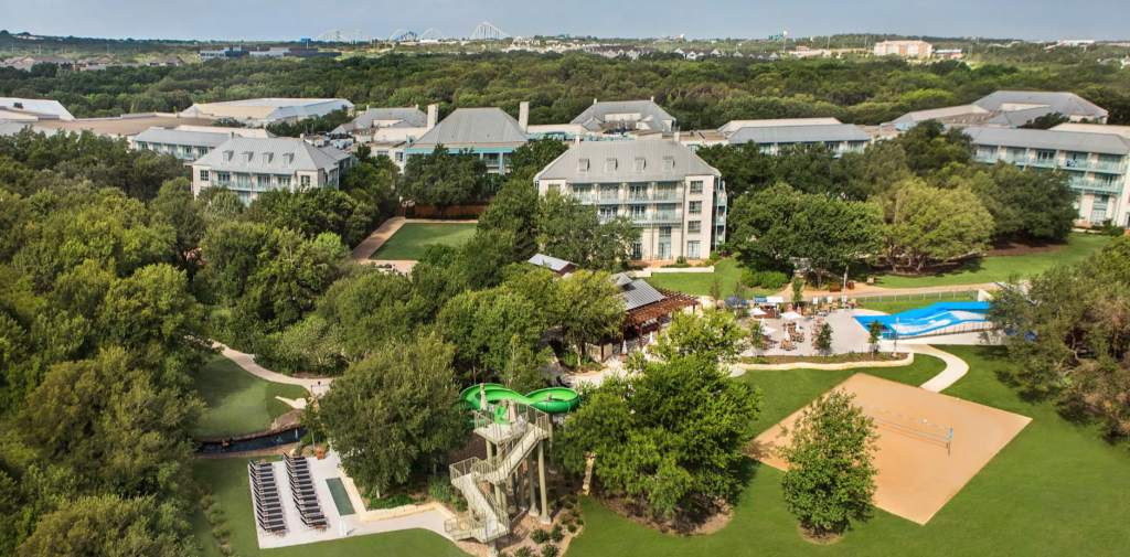 Aerial view of a resort-like campus with multiple gray-roofed buildings surrounded by lush trees and green lawns, with a pool area and water slides in the foreground.